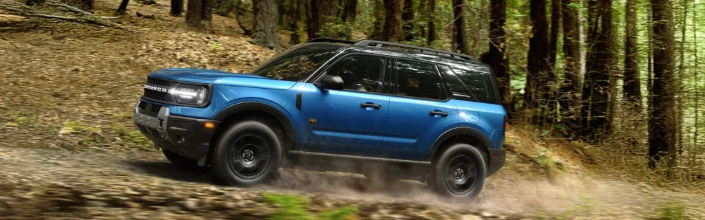 The image shows a blue Ford Bronco Sport SUV driving through a forested, off-road trail.